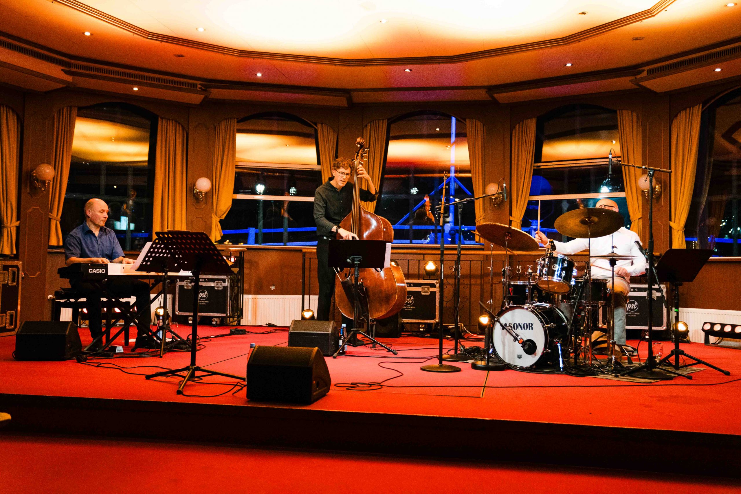 Jazz-Trio live auf der Winter Jazz Cruise in Hamburg: Pianist, Kontrabassist und Schlagzeuger auf der Bühne der MS Louisiana Star, festliche Atmosphäre mit Blick auf den beleuchteten Hafen.